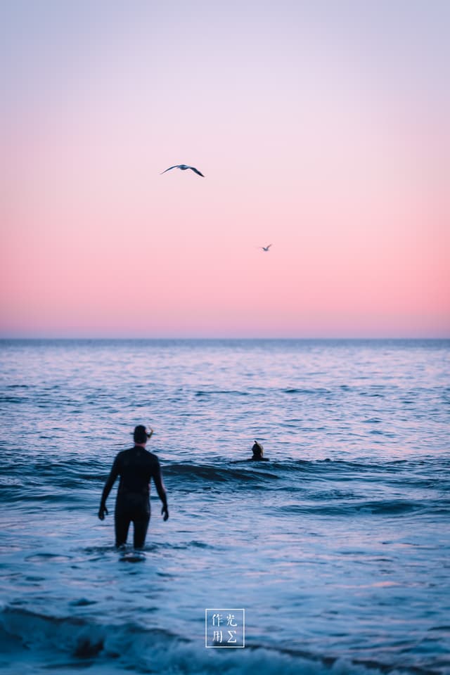 Wading into a pastel-drenched sea, a lone surfer’s silhouette cuts against a cotton-candy sky, board tucked under one arm as dusk’s pink and lavender hues melt into the horizon. Two distant figures bob on gentle swells, while seabirds wheel overhead, adding a whisper of motion to the tranquil expanse. The scene hums with quiet anticipation—a fleeting moment of calm before the next wave ride