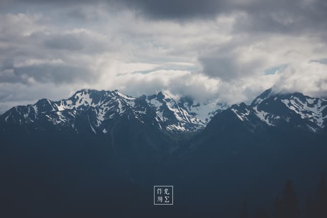 Jagged summits rake the sky, their snow-scalloped ridges etched in charcoal against roiling, slate-gray clouds. Pockets of silver light break through the storm-laden vault, tracing sharp contours and plunging the valleys into deep cobalt shadow. An electric hush grips the scene, as though the mountains themselves are holding their breath before the tempest unleashes