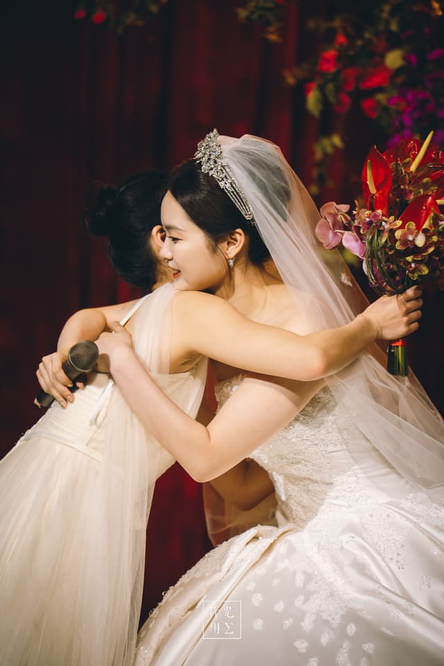 Clasping each other in a joyful embrace, the bride in a pearl-embroidered ball gown crowns the moment with a glittering tiara and sheer veil, while her friend’s off-shoulder chiffon dress ripples softly beneath warm chandelier light. A cascade of magenta orchids and crimson blooms spills from the bridal bouquet, punctuating the rich burgundy drapery with jewel-like intensity. Their radiant smiles and entwined hands transform lace and petals into a vibrant tapestry of love and celebration