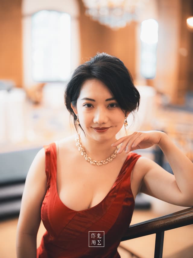 Glowing under the crystal chandelier’s golden haze, she perches with effortless poise—her ruby-red gown sculpting around graceful shoulders and drawing the eye to a delicate collarbone A lustrous gold necklace and matching earrings catch stray light, casting flickers across smooth skin Dark waves frame a calm, knowing smile, while soft-focus columns and opulent furnishings blur into a warm, amber backdrop The intimate interplay of light and shadow bathes every curve and contour in gentle drama, evoking a timeless elegance that lingers like a whispered promise