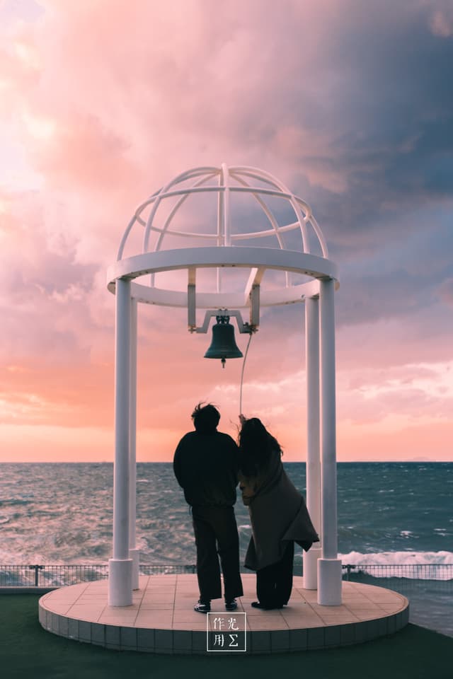 Bathed in a rose-and-lavender glow, a minimalist white pavilion crowns the shoreline, its single bell suspended above two dark silhouettes leaning into each other. Footprints trail through damp sand toward the gently lapping sea, while cotton-candy clouds drift across the horizon. The hushed rhythm of waves and the lingering warmth of dusk infuse the scene with intimate serenity