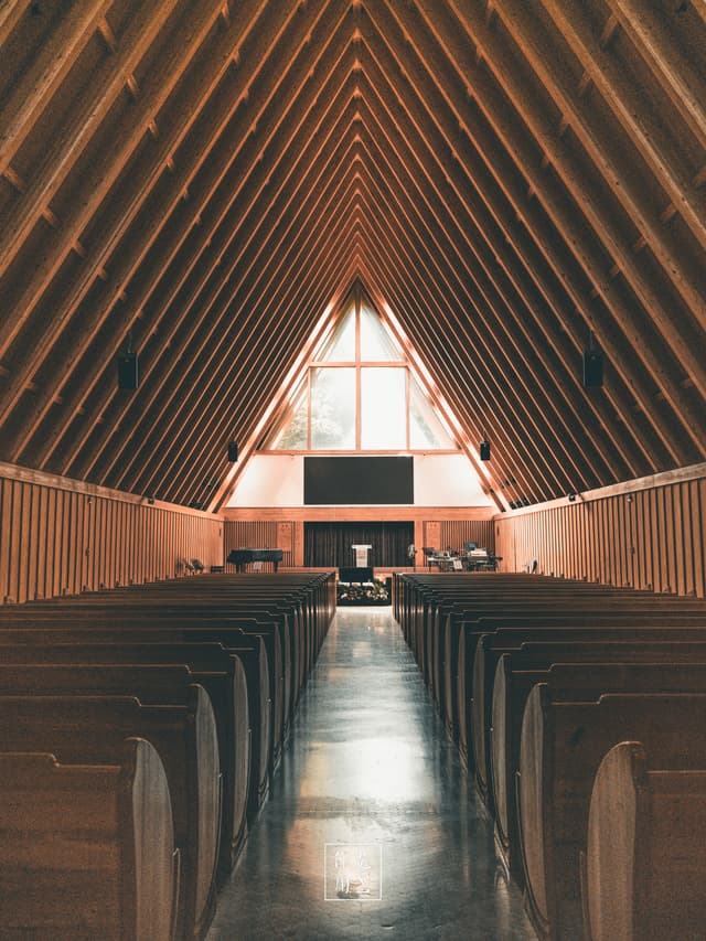 Sunlight streams through the soaring triangular window, casting a warm amber glow along the polished aisle. Slender wooden beams rise in perfect symmetry, converging overhead like silent sentinels. Rows of simple, curved pews flank the center walkway, their smooth grain accentuated by the soft, diffused light. At the far end, a modest altar bathed in gentle radiance anchors the space, lending an air of hushed reverence. The hush between the rafters and the golden hue creates a calm, almost meditative atmosphere—an architectural hymn in wood and light