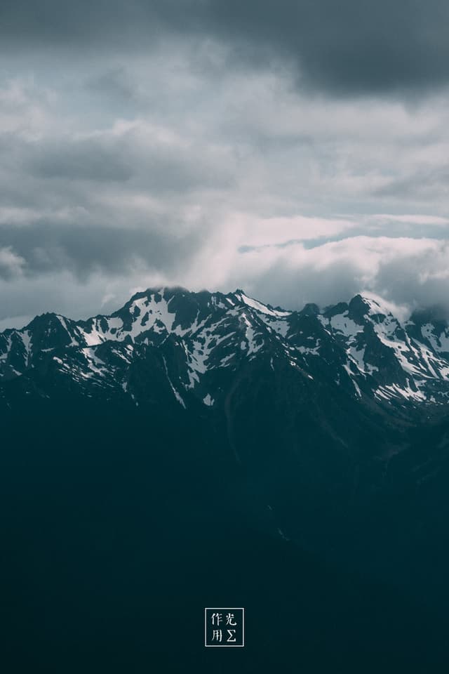 Jagged peaks thrust skyward, their dark flanks streaked with filigrees of snow that gleam like molten silver against bruised clouds. A heavy, brooding sky churns above, the low-hanging storm clouds parting in places to spill pale shafts of light onto the craggy ridges below. Mist clings to the mountain’s shoulders, softening the sharp silhouettes and lending a sense of shifting scale—as if the peaks might vanish back into the clouds at any moment. The overall palette of slate blues and steely grays deepens the mood, turning the scene into a raw, elemental tableau of rock, ice, and brewing tempest