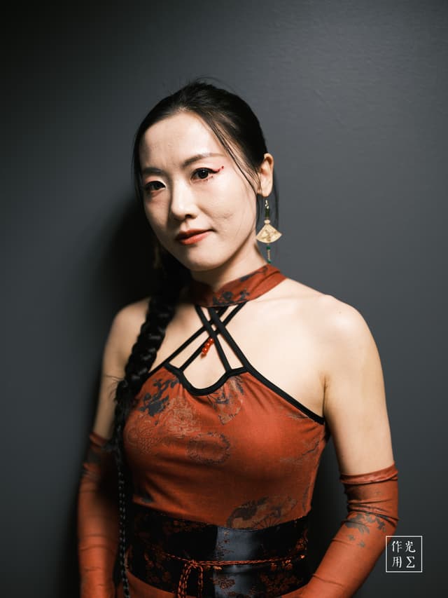 Amber light caresses her porcelain skin, revealing the intricate lattice straps and muted floral motifs of her deep-red cheongsam–inspired dress. A single braid drapes over one shoulder, its dark sheen contrasting the smooth charcoal backdrop. Her steady gaze and subtle, golden earrings catch the light just so, infusing the portrait with a quiet confidence that fuses timeless elegance and modern edge