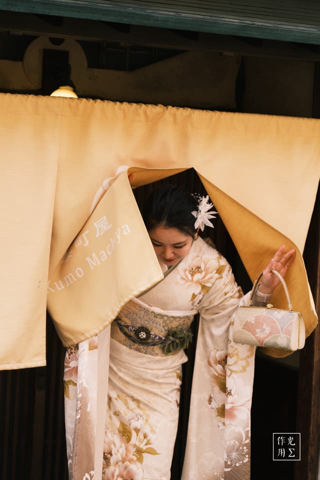 Sliding apart the ochre noren, she emerges swathed in an ivory kimono patterned with pale chrysanthemums, the silk’s soft sheen catching every flicker of the lantern light. Her obi’s intricate weave cinches the robes at the waist, while a single white blossom graces her dark hair, lending a whisper of spring to the warm, hushed entryway. The amber glow and tender tension in her downward gaze conjure a moment suspended between tradition and quiet anticipation