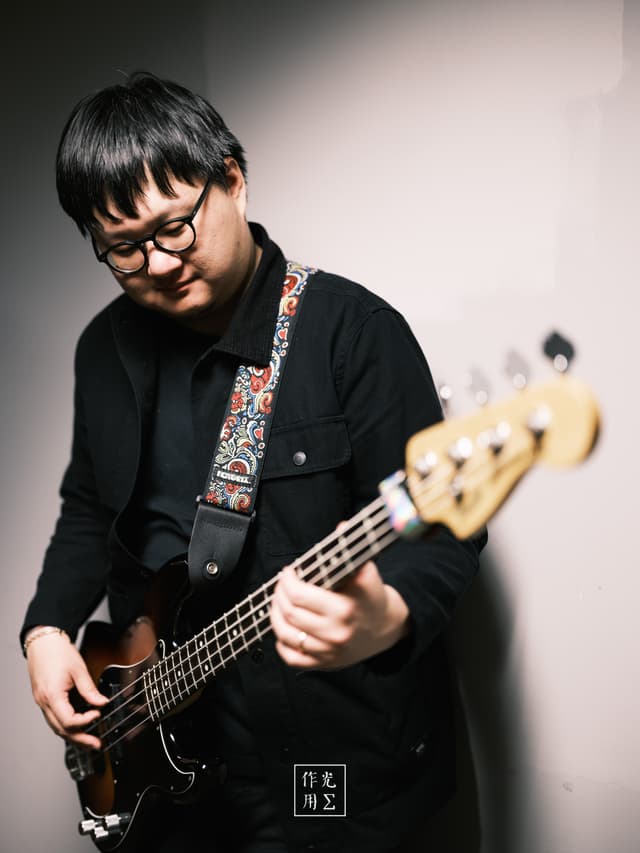 Fingers glide over the bass’s polished fretboard, coaxing warm, resonant tones beneath a soft, focused glow. A pop of paisley on the strap and the player’s quiet grin punctuate the deep shadows, while the blurred backdrop suggests an intimate, almost secretive rehearsal space. The interplay of sharp detail and dreamy bokeh turns this moment of concentration into a hushed celebration of sound and solitude