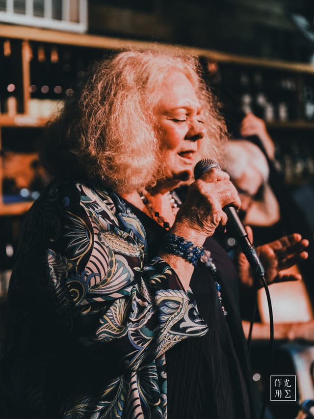 Silver-gray curls spill over her shoulders as she leans into the microphone, every wrinkle lit by the amber glow of the bar’s backlit bottles. A flowing kimono-style jacket, patterned with ivory ferns against midnight blue, ripples with each soulful phrase. Her eyes half-closed, she pours grit and grace into every note, the low hum of a bass and the clink of glasses weaving around her voice. Smoke-tinged air and warm wood paneling cradle the moment, giving her performance an intimate, almost sacred hush