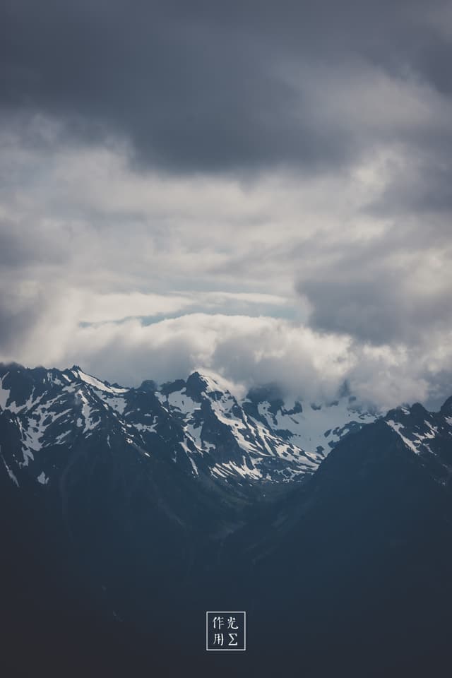 Jagged peaks thrust skyward, their snow-laced ridges cleaving through a churn of gunmetal clouds. A slender shaft of pale light seeps between storm-heavy layers, grazing icy summits and casting brooding shadows into the valley below. Wisps of mist coil around the mountains’ shoulders, heightening the raw, elemental drama of a landscape caught between light and looming tempest