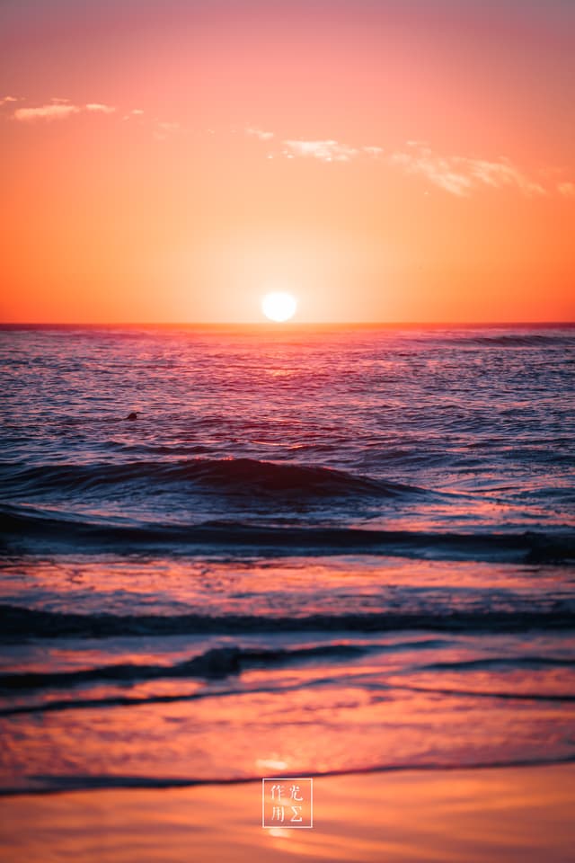 Sunlight spills like molten gold across a gently rippling sea, each wave tipped in rose and ember as the sun sinks toward the horizon. A soft haze of coral and lavender stretches overhead, blurring the boundary between sky and water. Wisps of cloud catch the last glow, their edges glowing neon against the deepening dusk. At the shoreline, translucent waves unfurl in rhythmic whispers, leaving wet sand shimmering with every hue of the fading day. A delicate artist’s seal in white punctuates the lower center, grounding this fleeting moment in timeless calm