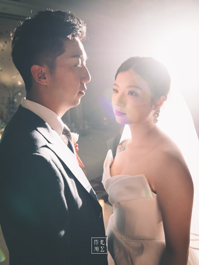 Bathed in a glowing halo of backlight, the groom’s sharp tuxedo contrasts with the bride’s sculpted white gown, its origami-like folds catching stray rays. Their faces hover close—his profile resolute, hers luminous and dream-wide—while a soft lens flare drifts between them, imbuing the moment with an otherworldly warmth. Dust motes drift through the light, freezing in midair like fairy dust, and the couple’s silent exchange pulses with tender anticipation