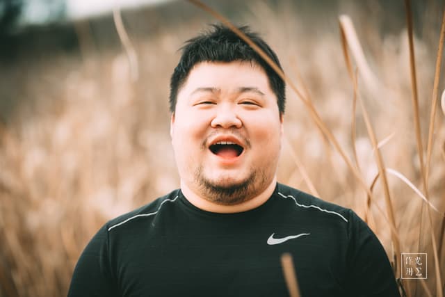 Beaming with unrestrained laughter, the man’s face lights up against a backdrop of golden, swaying reeds. Warm afternoon light softens the scene, brushing his features in a gentle glow that contrasts with the deep black of his Nike shirt. Every strand of grass around him seems to lean in, echoing the infectious joy that radiates from his wide-open mouth and sparkling eyes. The shallow depth of field blurs the horizon into a creamy wash of browns and tans, placing his exuberant presence sharply in focus and turning a simple moment into an unforgettable burst of warmth