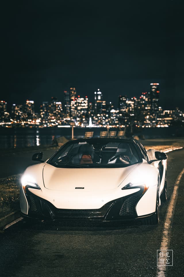 Glancing steel-white curves hug the pavement as twin LED “eyes” slice through the midnight haze. Gleaming carbon accents catch stray city sparks, while ripples of harbor light dance across the hood. Behind the poised McLaren, a constellation of skyscraper windows shimmers on dark water, lending the scene an electric tension—sleek power waiting in silent communion with the urban glow