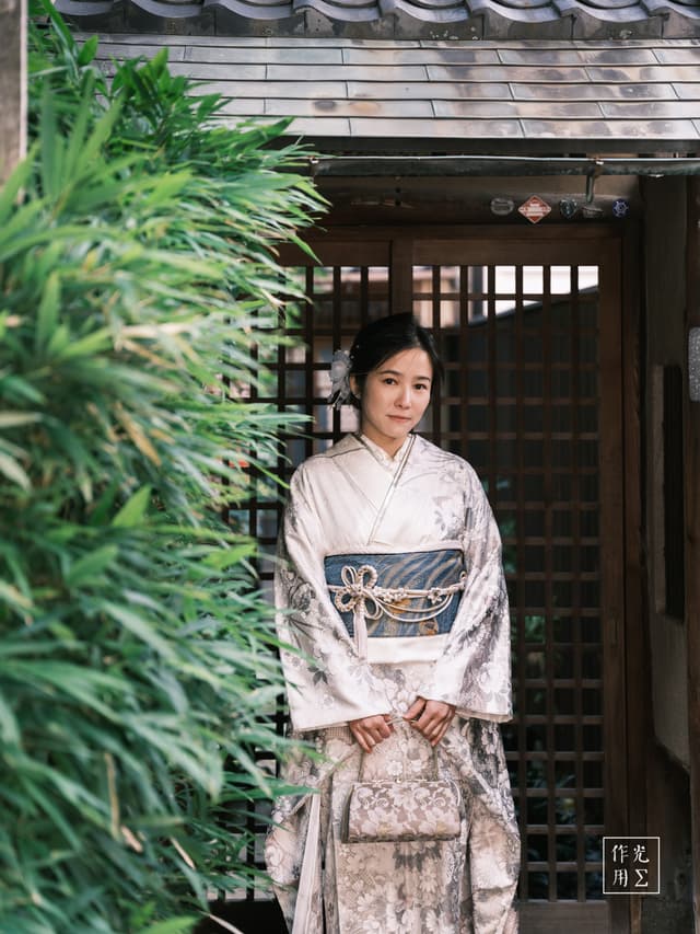 Soft daylight filters through the bamboo leaves, casting dappled emerald shadows across her pale silk kimono. She stands with measured grace before a weathered wooden lattice door, the floral embroidery on her sleeves whispering of spring blossoms long past. A slate-gray obi, knotted into a sculpted bow, cinches her waist, while a delicate brocade purse swings gently at her side. Quiet confidence radiates from her calm posture and the faint curve of her lips, as though she’s already slipped into a half-remembered dream of old Kyoto