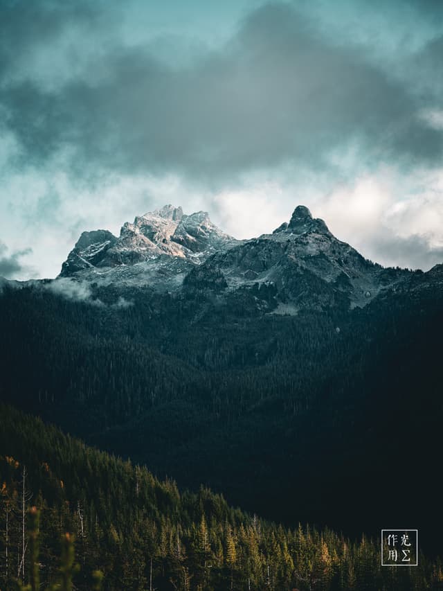 Cloaked in swirling mist, jagged granite spires thrust skyward, their snow-dusted summits glowing against a brooding, cobalt-tinged cloudbank. Beneath, the dark evergreen canopy stretches like a living carpet, its emerald tips catching the last glimmers of light. An electric tension hangs in the air, as if the mountains themselves are holding their breath before a storm