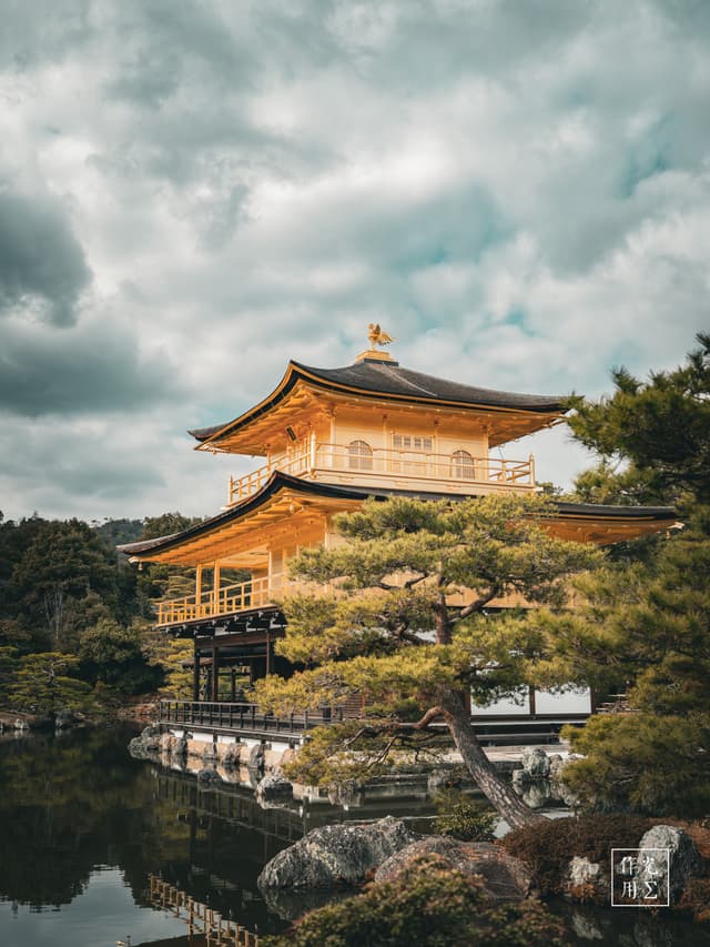 Gleaming golden tiers hover above a glass-smooth pond, their lacquered eaves catching the muted glow of an overcast sky. Gnarled pine branches frame the pavilion’s ornate silhouette, while ripples of emerald water mirror the temple’s gilded warmth against the brooding charcoal clouds. Moss-clad rocks and slender wooden walkways trace a serene path around the perimeter, inviting quiet reflection in the hush before an impending rain
