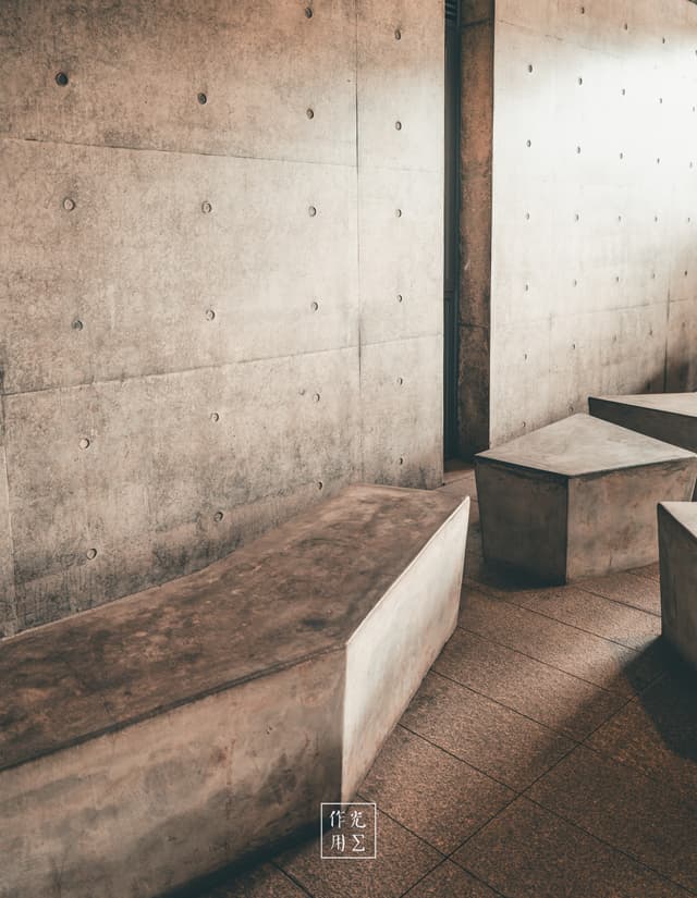 Battered concrete slabs rise and dip like monolithic waves across a muted tile floor, their sharp angles carving a rhythmic path along a raw, pock-marked wall A narrow shaft of light drapes across one block’s crisp edge, coaxing out the grainy texture and earthy warmth hidden in the cool gray The quiet interplay of shadow and form feels both austere and meditative—an impromptu altar to minimalist geometry, where every corner and surface whispers of stillness and restrained strength