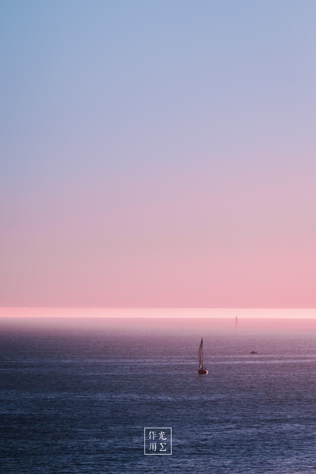 Drifting across a vast expanse of glassy water, a lone sailboat punctuates an ethereal horizon painted in pastel pinks and powder blues. Crisp horizontal bands of color blur sky and sea into one seamless canvas, while the tiny silhouette of the mast lends a whisper of human presence to the serene, almost otherworldly calm