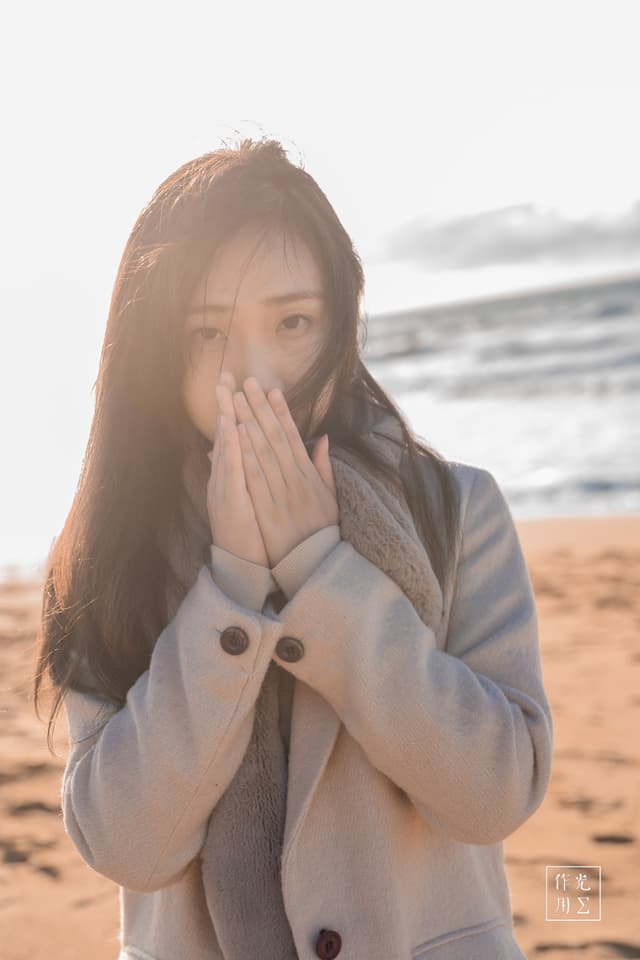 Bathed in a soft, golden-hour glow, she stands at the water’s edge in a tailored ivory coat, hands pressed to her lips against a brisk seaside breeze. Strands of dark hair drift across her face, catching flecks of sunlight like liquid amber. Behind her, gentle waves roll in under a pale sky, their muted turquoise and sand­stone tones echoing the calm yet brisk mood. The quiet tension between her protective posture and the vast, open shoreline creates an intimate, almost secret moment—where warmth and chill, light and shadow, meet in a single, breath-caught frame