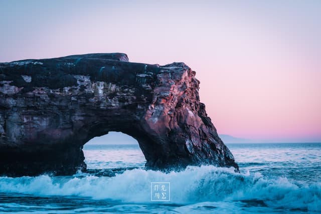 Beneath a pastel sky melting from lavender to blush, a rugged coastal arch juts into frothing teal surf. Golden-pink light bathes the limestone’s weathered contours, carving every crevice in warm relief. Rolling waves whisper through the opening, their foamy edges catching the sunset’s glow. The contrast of sharp stone against soft twilight sky lends the scene an otherworldly calm, as if time itself hesitates at this threshold between land, sea, and sky