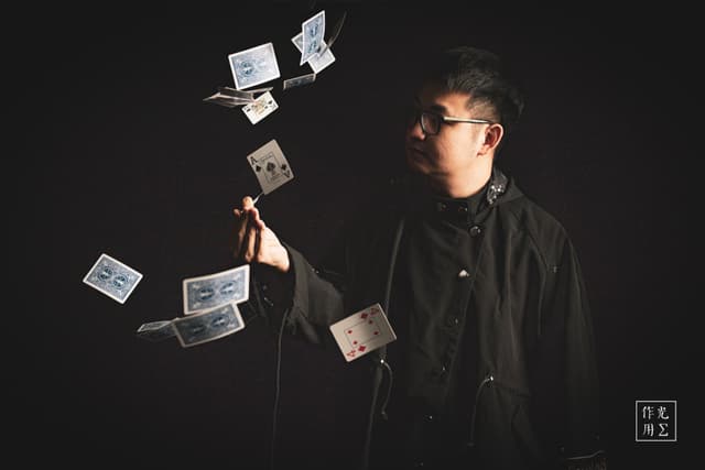Commanding the darkness with a single, outstretched wand, a young magician conjures a graceful spiral of playing cards that seem to float in mid-air. Sharp side lighting carves his focused profile and the crisp edges of each card against the inky black backdrop. His charcoal shirt and round glasses lend an aura of understated poise, while the cards’ white faces and bold pips shimmer like fragments of starlight. The scene pulses with quiet suspense, as if time itself has paused to watch this delicate dance of sleight and shadow