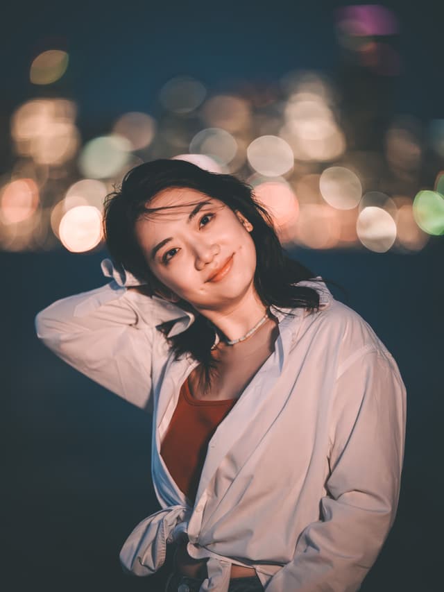 Bathed in the soft glow of city lights, she tilts her head just so, one hand drifting behind her neck as if to catch the night’s breeze Her dark hair frames a gentle smile, eyes sparkling against the blurred orbs of neon and streetlamps dancing in the distance A crisp white shirt draped over a rich, rust-red camisole adds a casual elegance, its loose tie at the waist whispering carefree confidence The warm bokeh backdrop pulses with energy, lending the scene an intimate yet electric atmosphere