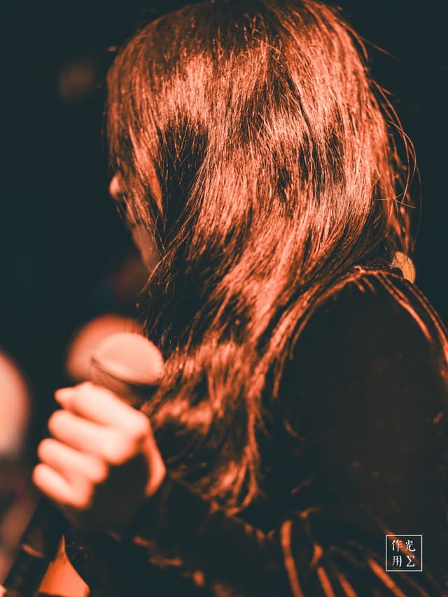 Cascading auburn waves glimmer under a warm spotlight, each strand catching the stage’s copper glow. A slender hand grips a vintage microphone, knuckles white against its polished metal. Velvet sleeves absorb the surrounding darkness, lending depth to the scene’s rich, sensuous palette. Hints of distant bokeh lights swirl behind her, as if the audience’s heartbeat has been distilled into glowing orbs. The air feels thick with anticipation, charged by the promise of a voice about to rise