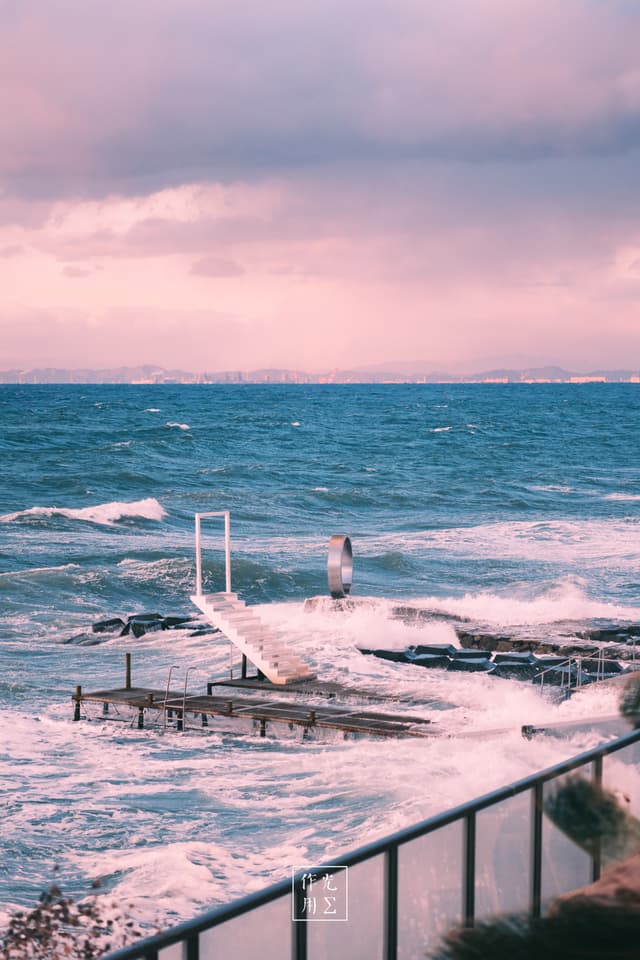 Surging foam lashes a weathered pier crowned by a lone diving ladder, its white railings streaked with salt. A restless cobalt sea churns beneath a blush-pink sky, clouds swirling in lavender and rose. Splintered planks dip beneath each wave’s crest while a wind-bent railing in the foreground frames the untamed dance of water and wood. Distant hills melt into the horizon’s rosy haze, lending a dreamlike cast to the raw, tempest-tossed energy
