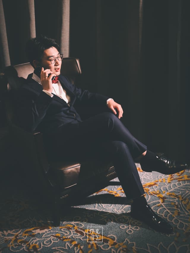 Leaning back in a deep leather armchair, he clasps a slim smartphone to his ear, the glow of its screen tracing the sharp line of his jaw His midnight-blue suit catches the soft lamplight, each fold and seam rendered in rich shadow and subtle sheen Behind him, heavy velvet drapes fall in brooding, cathedral-like folds, absorbing warmth and creating a cocoon of hushed secrecy Below, an ornate carpet patterned with gold and slate-blue flourishes cushions his polished Oxfords, whose gleam hints at fastidious care A faint haze drifts through the air, lending the scene a film-noir tension—as though every whispered word might unravel a high-stakes deal