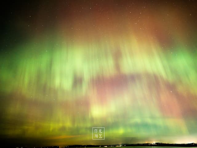 Draping the night sky in rippling ribbons of neon green and rose-pink, the aurora shimmers like celestial curtains caught between dancing waves and stillness Wisps of emerald light stretch downward in soft, vertical flares, while a faint, warm glow along the horizon hints at distant lanterns or the first blush of dawn The scene feels alive—ethereal, otherworldly, and poised on the edge of a dream