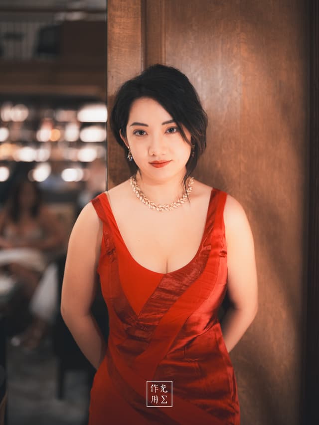 Bathed in golden bistro light, a woman in a crimson silk gown leans against a polished wooden column, the dress’s draped neckline catching every glint of illumination Her dark waves frame a calm, confident gaze, while a delicate diamond necklace sparkles at her throat In the softly blurred background, candlelit tables and murmured conversations lend the scene an intimate, almost cinematic hush