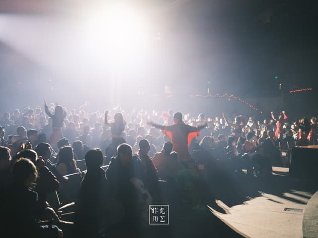 Jets of white light pierce through a haze of swirling dust, casting the crowd into sculpted silhouettes. A lone figure in a crimson shirt stands center stage, arms outstretched as if conducting the electric energy rippling through the arena. Faces blur into shadows and glimmering flecks in the air, each beam carving out momentary halos above waving hands. The atmosphere crackles with anticipation—raw, kinetic, and drenched in the promise of an unforgettable crescendo