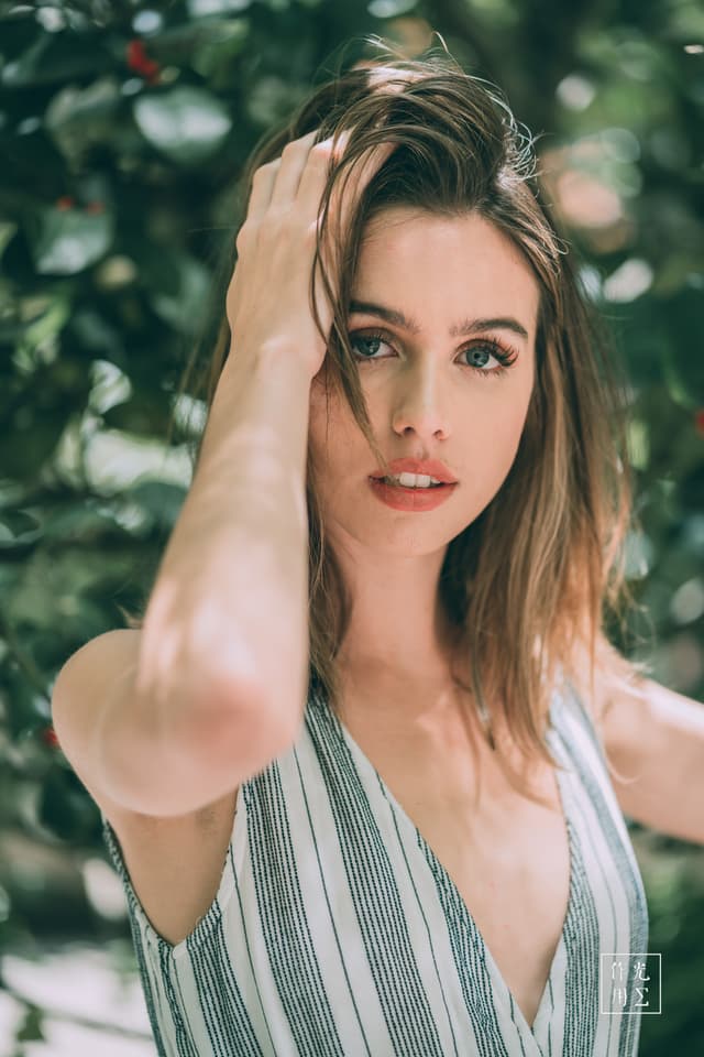 Sunlight filters through lush leaves, dappling her tousled chestnut hair and warm complexion. Slender fingers sweep strands aside, unveiling vivid green eyes that gleam with quiet confidence. Soft linen stripes in pale teal and ivory hug her shoulders, offering a breezy contrast to the deep foliage. The shallow depth of field blurs the surrounding greenery into a painterly haze, evoking the hush and mystery of a hidden garden