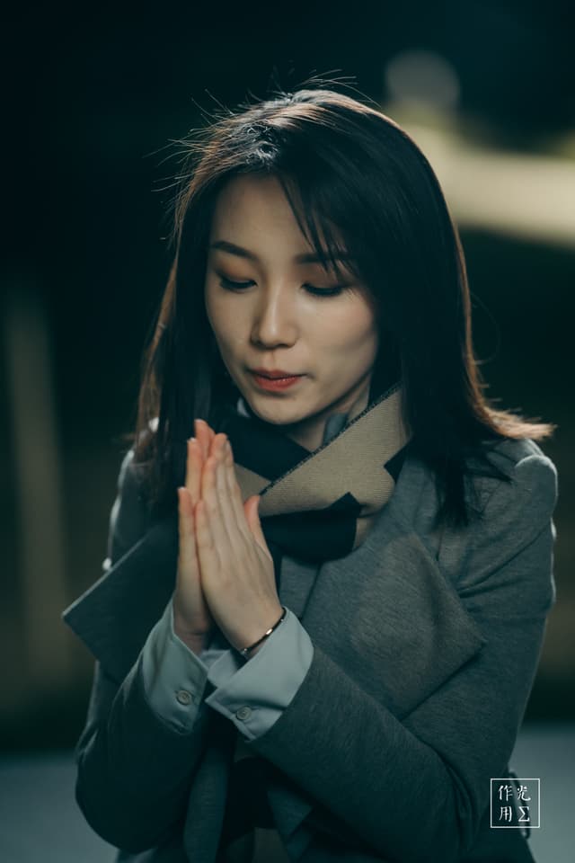 Warm lamplight spills across her face, casting gentle shadows that cradle her closed eyes and softly curved lips. Fingers pressed together glide upward, accentuating the quiet stillness in the bridge of her nose and the arch of her brows. A charcoal-gray coat and striped scarf wrap her in muted winter tones, while a blurred background of distant lights hints at a cold evening street. The contrast between the soft glow on her skin and the deep shadows around her creates an intimate moment of calm focus, as if time itself has paused around her silent prayer