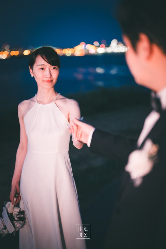 Soft halos of city lights dance across the river as she lifts her gaze, pale halter gown flowing like liquid moonlight His tuxedoed silhouette reaches toward her, offering a single white rose as bokeh orbs swirl behind them in gentle motion Her calm smile and lantern-soft skin catch every shimmer, while the night air brims with unspoken vows Cool ripples at their feet contrast with the warm glow on her cheeks, forging a tender harmony of light and shadow that feels both intimate and timeless