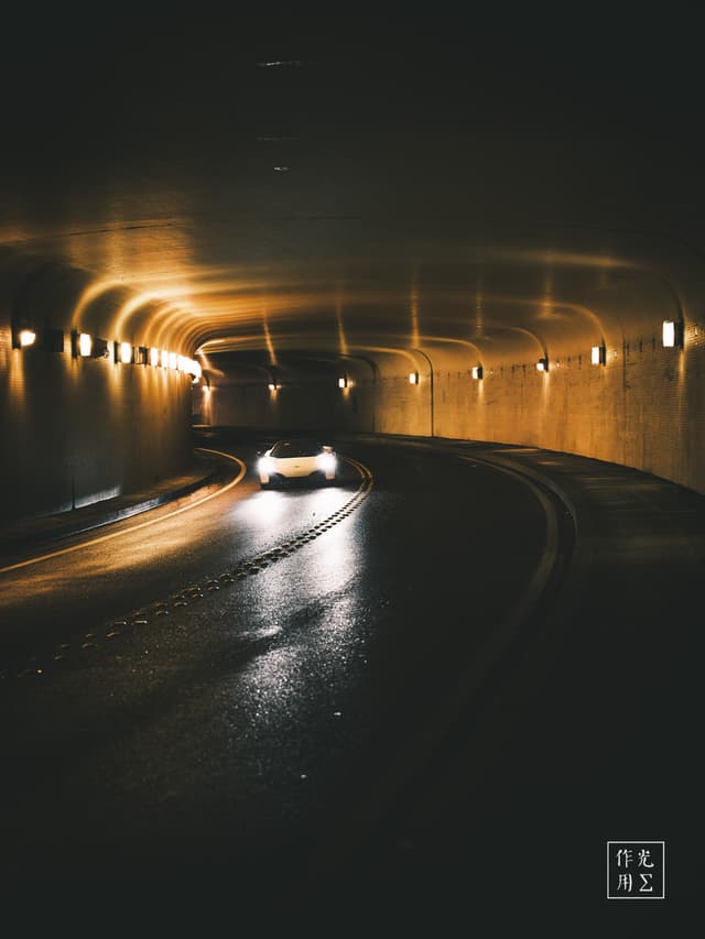 Glowing amber arches drip warmth across the glossy asphalt as a lone car glides through the curving tunnel. Soft pools of light ripple off the wet road, tracing the vehicle’s sleek silhouette and casting elongated shadows along the brick-lined walls. The tunnel’s rhythmic lamps create a hypnotic sequence of bright halos, guiding the eye toward the distant, inky exit. A whisper of motion blur lends the scene a breathless tension, marrying solitude and momentum under an almost cathedral-like ceiling of light. Each reflection and contour heightens the sense of passage through an otherworldly urban corridor