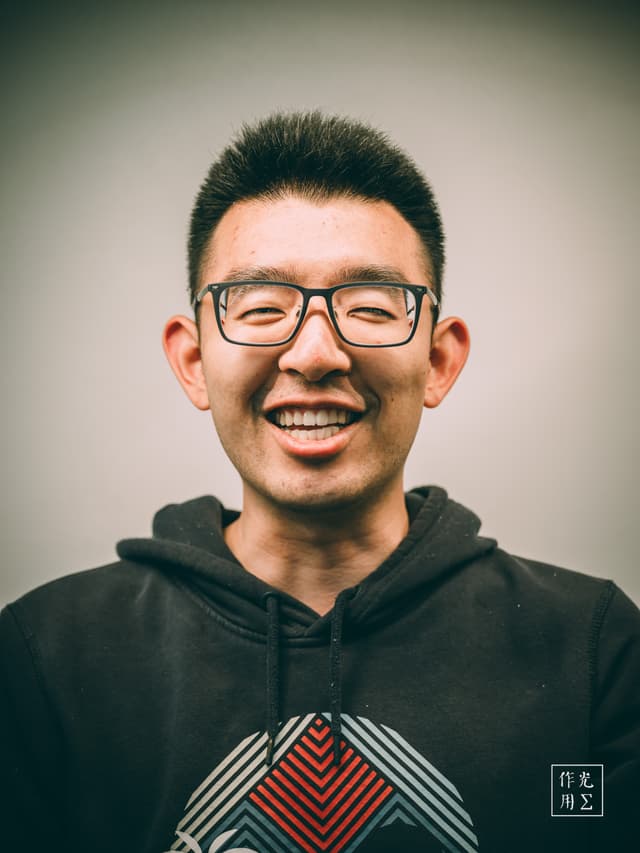 Soft amber light wraps around the subject’s face, casting gentle shadows that sculpt his high cheekbones and broad smile. Neat spikes of dark hair crown his forehead, while rectangular glasses catch subtle glints of reflection. His eyes crinkle with genuine warmth, illuminating a moment of unguarded joy. The deep-charcoal hoodie drapes casually over his shoulders, its bold geometric emblem at the chest lending a modern, urban edge. A faint vignette frames the portrait, drawing focus inward and heightening the sense of intimacy. The muted background gradient fades from sage to cream, amplifying his presence without distraction. In the lower corner, a discreet seal of characters hints at artistic intent, making this portrait not just a snapshot but a crafted encounter between subject and viewer