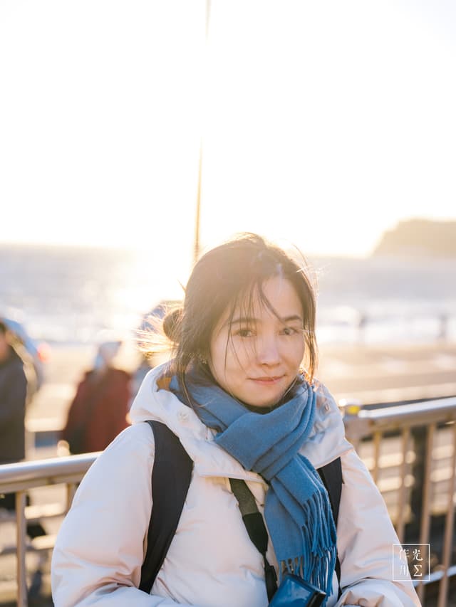Bathed in golden-hour glow, her sapphire scarf and snow-white coat pop against the chilly sea breeze. Wisps of hair catch the sun’s rays, framing a quiet smile as the coastline melts into soft-focus silhouettes beyond the railing. The interplay of warm light and crisp winter air gives the moment a serene, almost ethereal glow