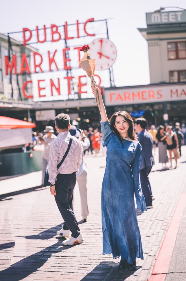 Sunlight slants across the red-brick pavement, illuminating a young woman in a flowing cobalt dress as she strikes an exuberant pose, one arm pointing straight up at the giant neon clock and “Public Market Center” sign overhead Shadows of midday bustle stretch behind her—tourists milling past colorful stalls, farmers’ market banners fluttering in a gentle breeze Her radiant smile and the vibrant blue of her dress pop against the vintage market façade, capturing a moment of spontaneous joy amid the timeless hum of urban life