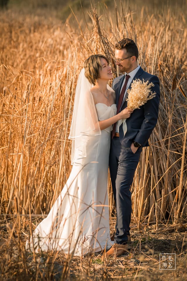 Sunlight filters through towering, amber-hued reeds as the bride’s ivory gown glides against the groom’s navy suit, their silhouettes softened by the golden hour glow. The bride’s delicate lace bodice and flowing veil contrast sharply with the groom’s sharp lines and textured tie, creating a harmonious balance of elegance and formality. Wisps of dried grasses in her bouquet echo the surrounding stalks, weaving the couple seamlessly into the autumnal landscape. A gentle breeze stirs the reeds, lending the moment a hushed intimacy and timeless romance