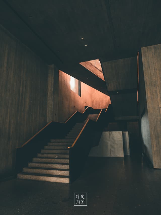 Carved from raw concrete, the broad staircase climbs into a narrow sliver of warm light, its sharp angles slicing through deep shadows Polished handrails catch a copper glow where the sun breaches the rigid geometry, turning austere walls into an almost living landscape of light and dark Above, a tilted ceiling panel paints a soft amber streak across heavy pillars, while the silent emptiness of the space feels both foreboding and serene Every surface exudes brutalist austerity, yet the sudden burst of sunlight injects a quiet drama that transforms cold structure into poetic architecture