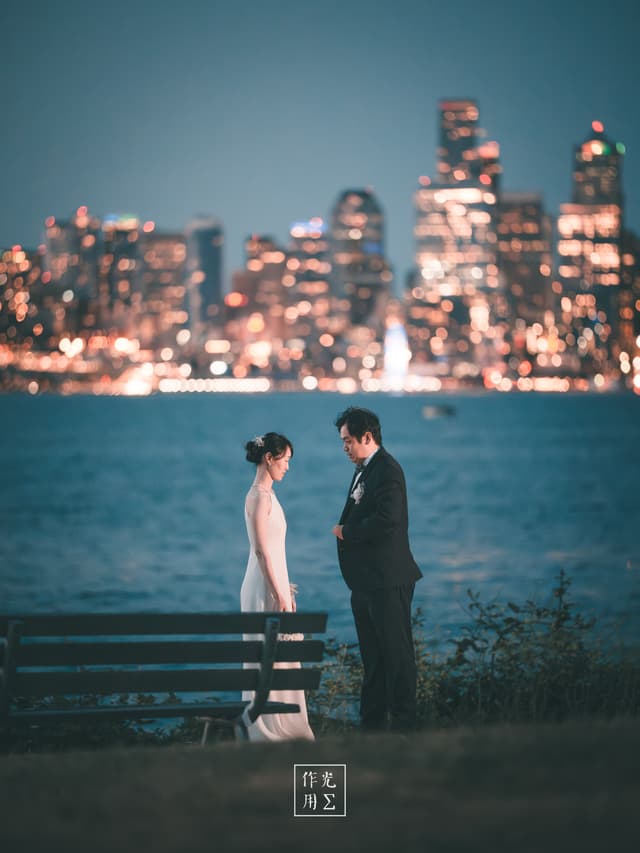 Glistening city lights ripple across the water as a couple stands at the water’s edge, the woman in a sleek ivory gown and the man in a tailored black suit A lone bench anchors the foreground, its empty slats echoing the quiet anticipation between them Behind, towering skyscrapers blur into a sea of gold and teal bokeh, turning the skyline into a dreamy, cinematic backdrop Cool night air drifts in off the bay, carrying hushed warmth of their whispered words and the faint hum of distant traffic Every glow and shadow frames their shared moment, balancing urban energy with intimate stillness