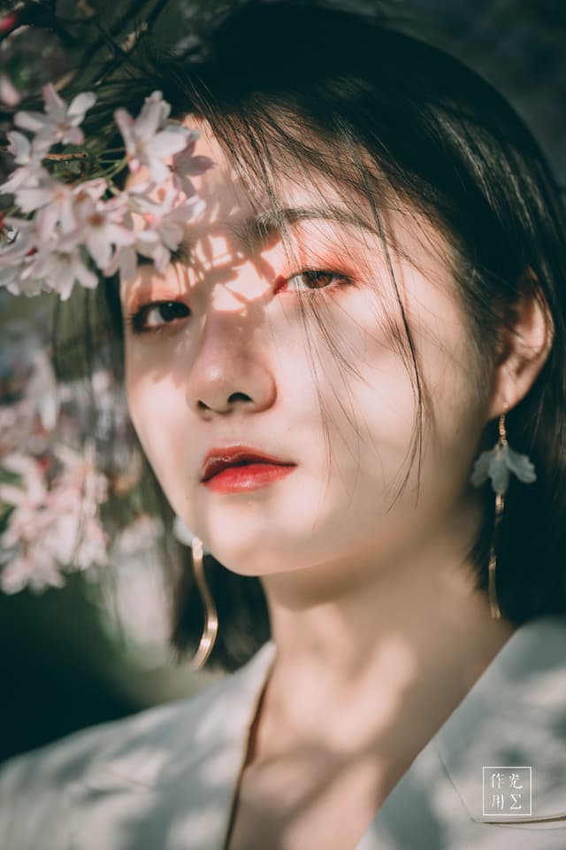 Soft sunlight dances through pale petals, casting lace-like shadows across her porcelain skin. Petal clusters hover at her temple, their soft pinks echoing the flush on her lips and the warmth in her dark, contemplative eyes. Loose strands of hair catch the light, framing her face with effortless grace, while delicate earrings sway just beyond the reach of blossom. The scene breathes a hushed springtime poetry—fleeting, fragile, and achingly alive