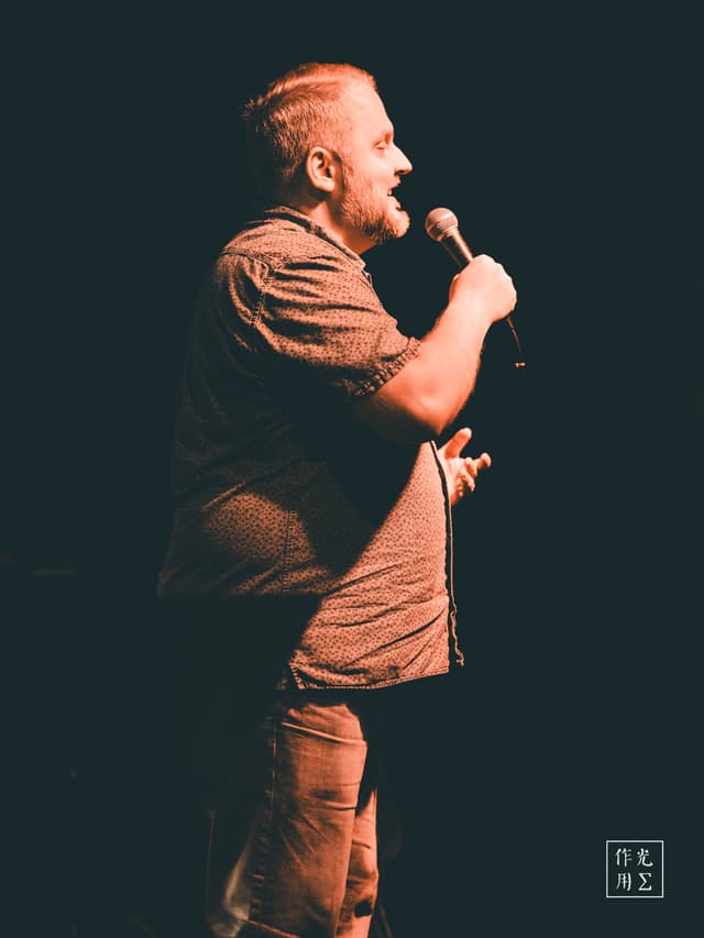 Bathed in a single warm spotlight, a bearded man leans into the microphone, every thread of his patterned shirt and ripple of his jeans sharply etched against the shadowed void behind him. His right hand grips the mic stand with quiet intensity while his left drifts outward, as if daring the silence to respond. Deep ochre highlights trace the curve of his jaw and the folds of fabric, turning an ordinary stage into a private theater of raw presence and hushed anticipation