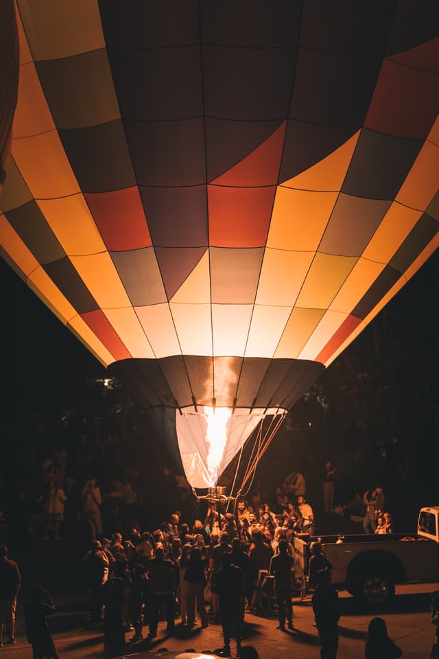 Flickering flames ignite the base of a striped hot-air balloon, spilling molten gold and amber across upturned faces. The checkerboard canopy towers overhead, its warm hues bleeding into the velvet night. Silhouetted figures cluster around the basket, their outlines quivering in the ember glow as heat ripples skyward. A low hum of anticipation vibrates through the air, thick with the scent of propane and whispered awe