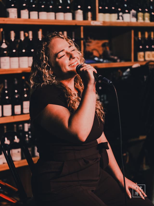 Bathed in amber light, a curly-haired vocalist tilts her head back, eyes closed, pouring raw emotion into every note. Her hand grips the vintage microphone as golden highlights dance across her face, accentuating the curve of her smile mid-crescendo. Rows of wine bottles stacked behind her blur into warm hues, framing the scene with an intimate bistro charm. The dim, cozy atmosphere hums with hushed anticipation, each listener drawn into the magnetic pull of her soulful performance