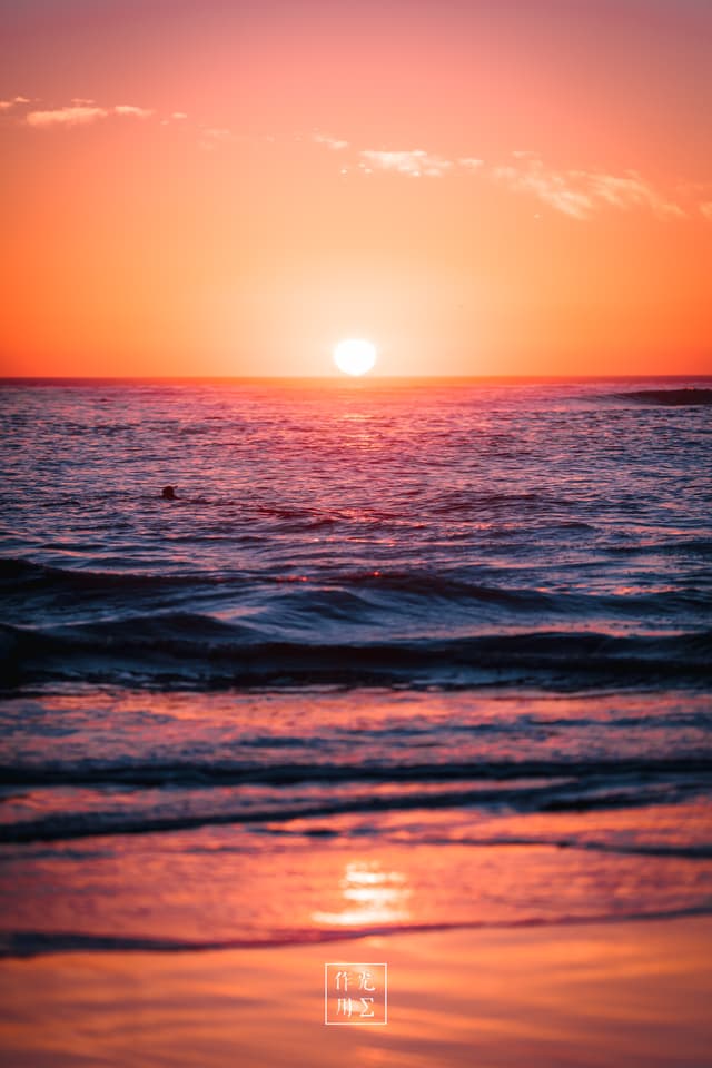 Molten gold spills across violet-tinged waves, each ripple catching fiery streaks as the sun hovers just above the horizon. Wisps of cloud drift like ember-dusted veils, softening the blaze into a peach-pink glow. A cool breeze stirs the salt-kissed air, carrying the whisper of the endless deep. The shoreline shimmers in mirrored light, where water and sky blur into a single, hushed moment of quiet radiance