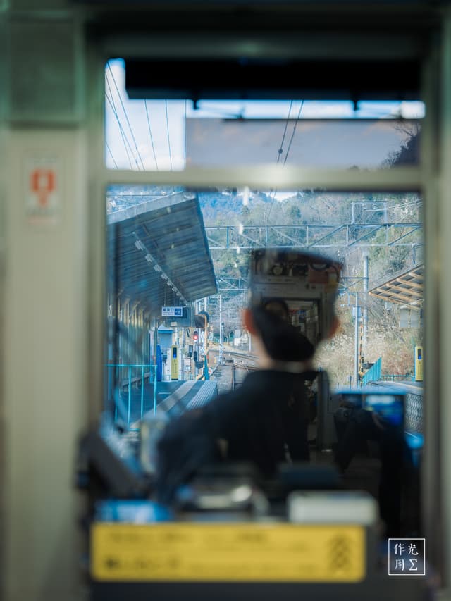 Sunlight filters through the driver’s cabin, illuminating the throttle and gauges with a warm glow as the operator stands poised at dawn. His dark silhouette bisects a mosaic of steel girders and crisscrossed catenary wires beyond the glass, the station platform’s edges softened into gentle blur. An electric hum lingers in the air, mingling with distant birdsong from the embankment—an atmosphere charged with quiet anticipation and the promise of motion