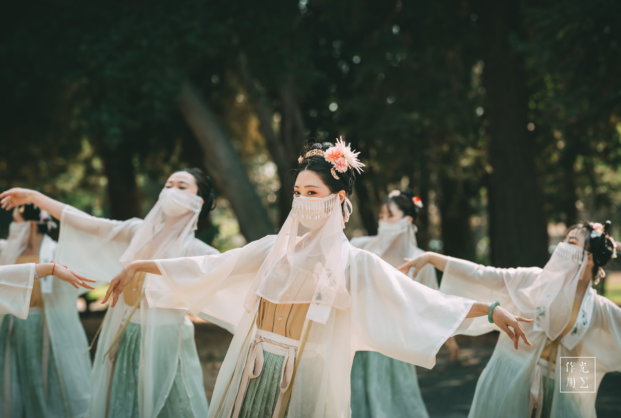 Swirling gauzy robes in mint and ivory drift like spring mist through a sun-dappled grove. Veiled dancers extend slender arms in seamless harmony, silk sleeves rippling with each measured step. Delicate peony crowns perch atop soft chignons, their pastel blooms echoing the pale green of the flowing skirts. Slanted beams of light pierce the oak canopy, setting airborne dust motes aglow and bathing the troupe in an almost otherworldly hush. Each graceful movement feels like a whispered invocation of dawn amidst ancient trees