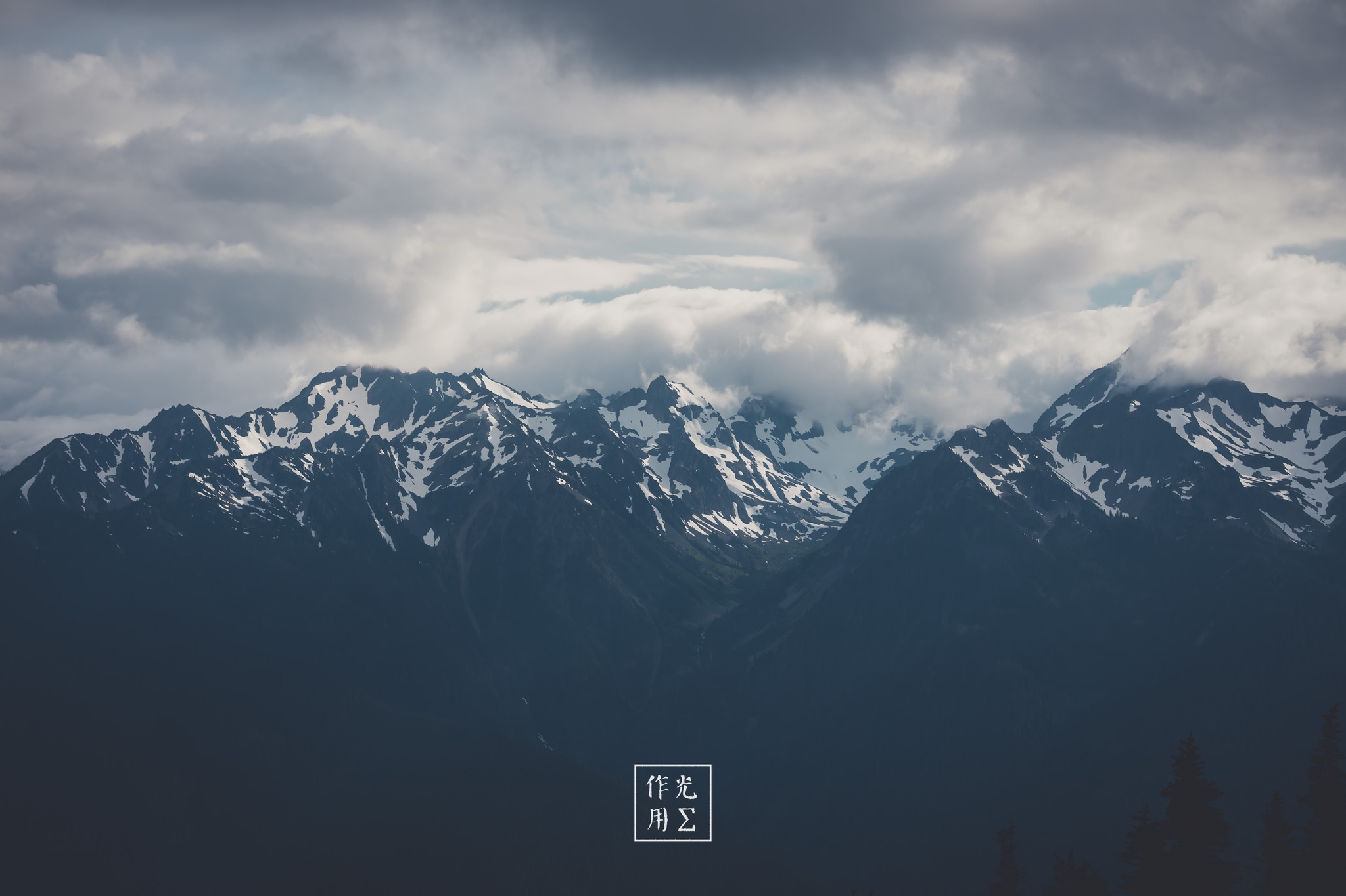 Jagged summits rake the sky, their snow-scalloped ridges etched in charcoal against roiling, slate-gray clouds. Pockets of silver light break through the storm-laden vault, tracing sharp contours and plunging the valleys into deep cobalt shadow. An electric hush grips the scene, as though the mountains themselves are holding their breath before the tempest unleashes