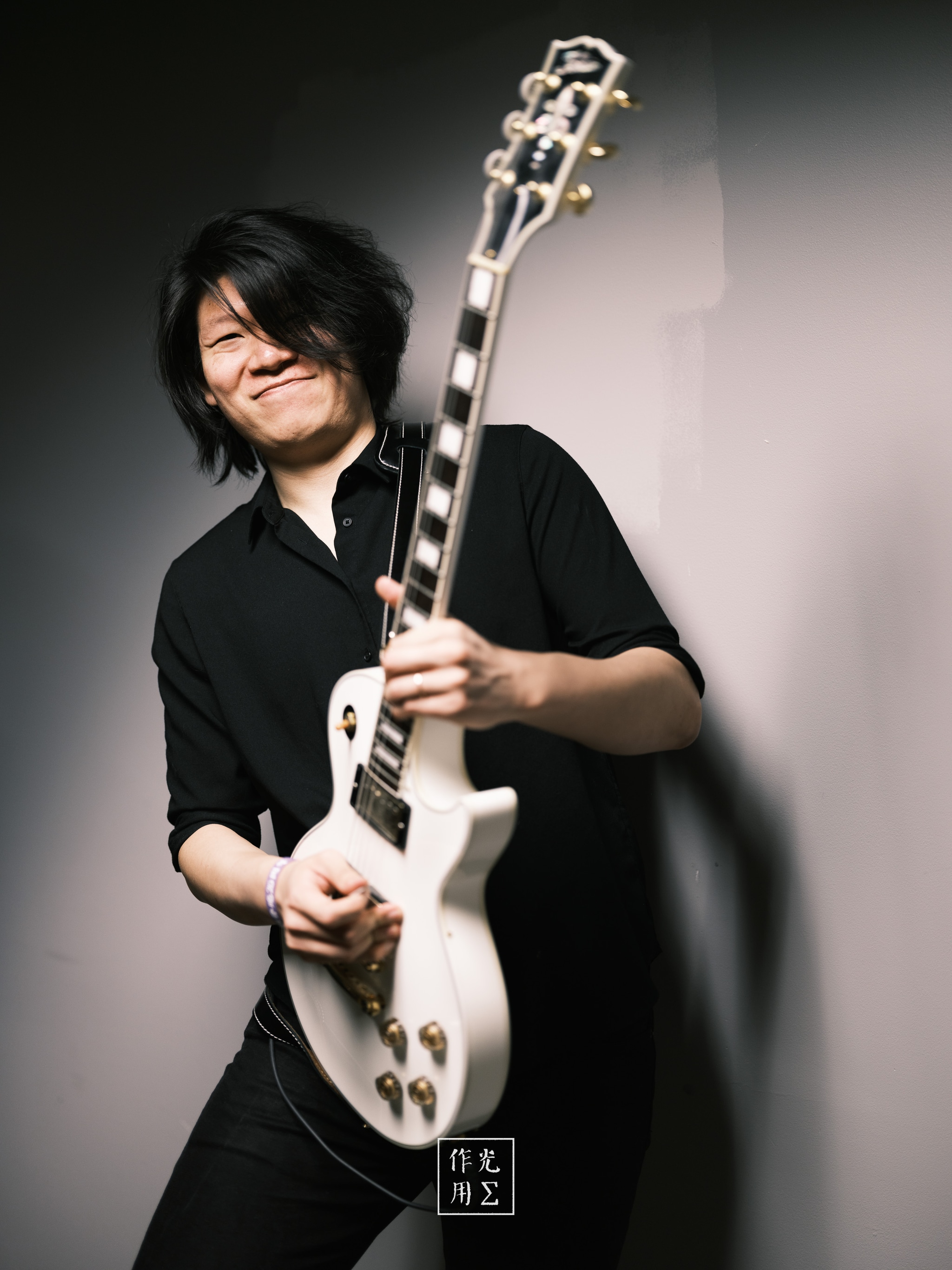 Cradling a snow-white Les Paul like a long-lost confidant, the guitarist’s midnight-black shirt and raven hair pop against the soft, gradient backdrop. A single beam of light carves sharp edges along the guitar’s curves, casting elongated shadows that dance across the wall. His easy grin and relaxed stance inject the scene with playful confidence, while the gleaming gold hardware and polished fretboard lend an air of meticulous craftsmanship. Together, instrument and player strike a bold, minimalist tableau—where every highlight and shadow whispers rock-steady cool